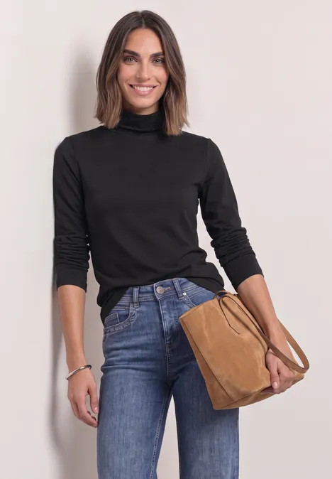 Eine lächelnde Frau in schwarzem Rollkragenpullover und Jeans, hält eine braune Handtasche.