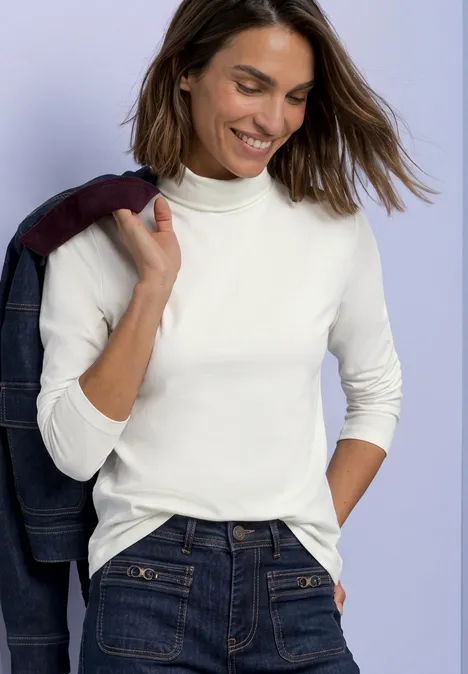 Femme souriante en pull à col roulé blanc et jean, portant une veste sur l’épaule devant un fond violet.