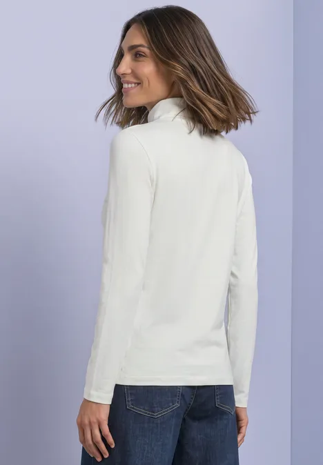 Une femme souriante en pull à col roulé blanc regarde par-dessus son épaule sur fond violet.