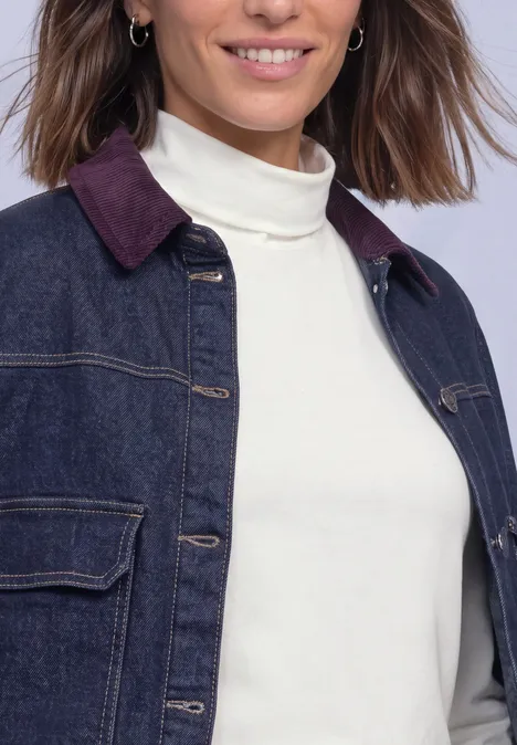 Une femme porte une veste en jean foncé avec col bordeaux et un pull à col roulé blanc.