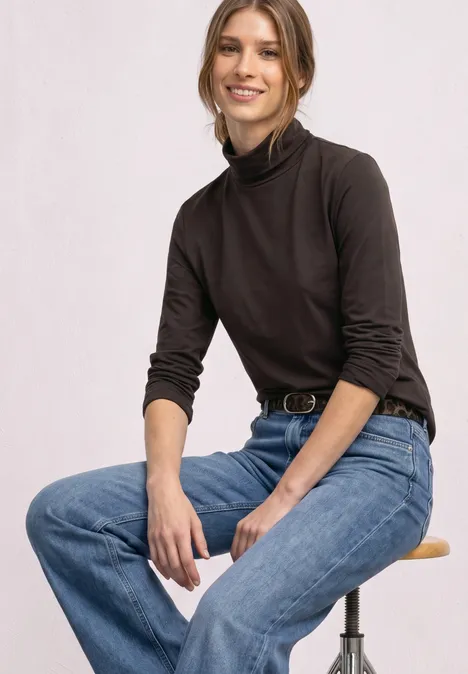 Eine lächelnde Frau in braunem Rollkragenpullover und blauen Jeans sitzt entspannt auf einem Hocker.