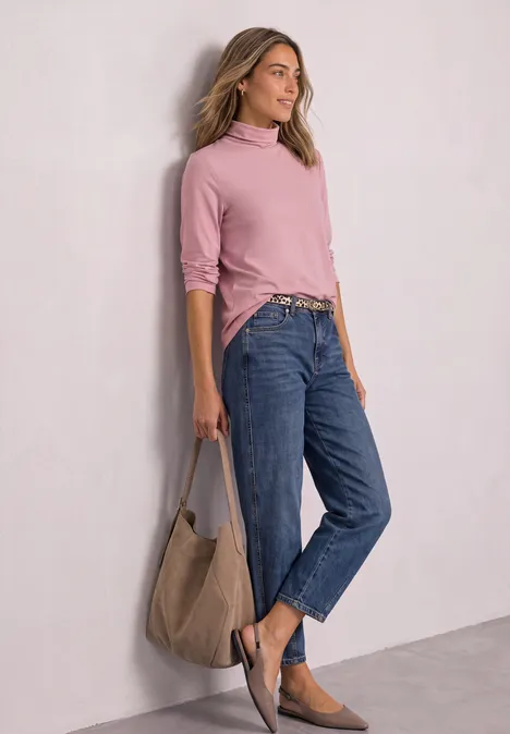 Une femme appuyée contre un mur porte un pull rose, un jean bleu, des mocassins en daim et un grand sac.