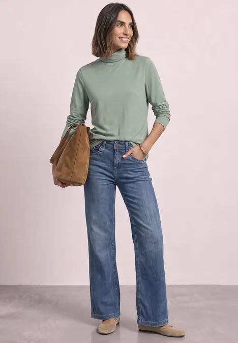 Langarm Shirt in Unifarbe mit Rollkragen eucalyptus green Frau mit grünem Rollkragenpullover, geraden blauen Jeans und brauner Tasche lächelt vor hellem Hintergrund.