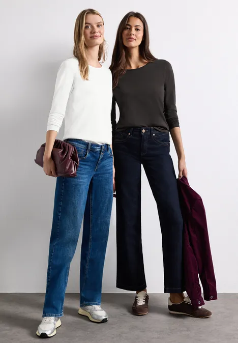 Zwei Frauen posieren in lässiger Alltagskleidung: helles Oberteil und Jeans, dunkler Pullover und weite Hosen.