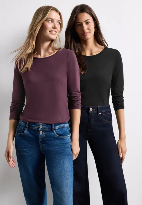 Deux femmes portent des hauts à manches longues et des jeans, posant côte à côte sur fond blanc.
