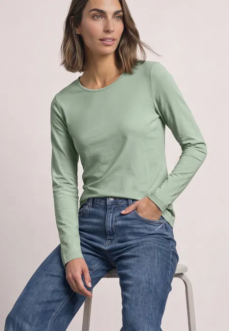 Eine Frau sitzt lässig auf einem Hocker, trägt ein hellgrünes Langarmshirt und blaue Jeans, eine Hand in der Tasche.