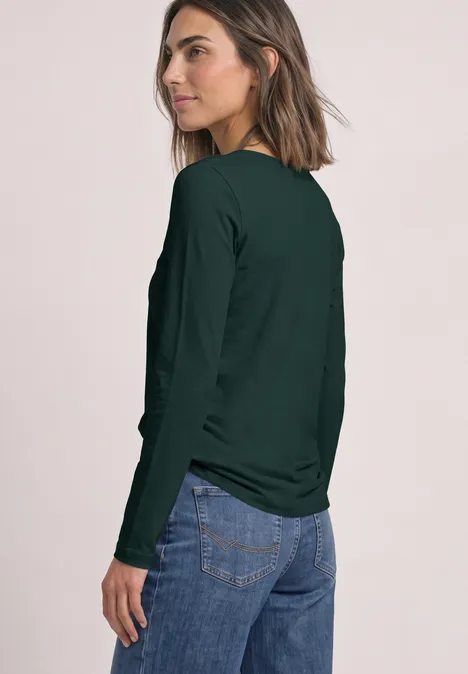 Frau trägt dunkelgrünes Langarmshirt und Jeans, blickt über die Schulter vor hellem Hintergrund.