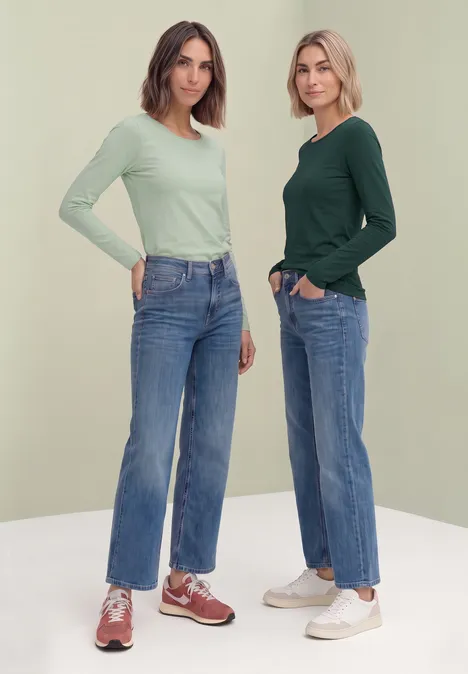 Zwei junge Frauen in Jeans, langärmligen Oberteilen und Sneakern vor hellem Hintergrund.