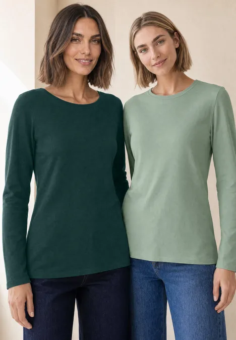 Twee vrouwen glimlachen in lange mouwt-shirts in groen tinten en jeans in een lichte setting.