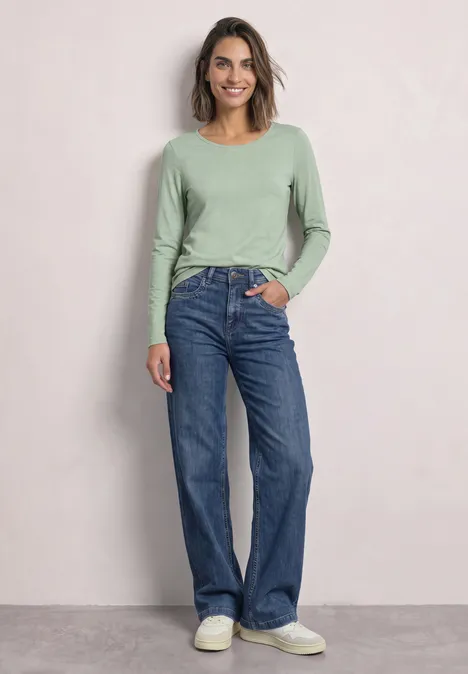 Eine Frau trägt ein grünes Langarmshirt, weite Jeans und weiße Schuhe vor hellem Hintergrund.