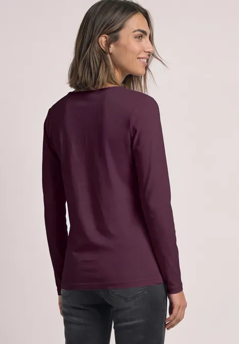 Frau von hinten in dunkellila Langarmshirt vor neutralem Hintergrund.
