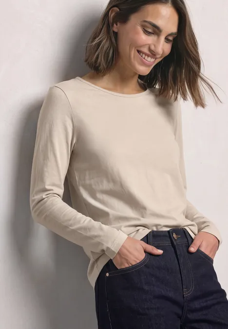 Frau lehnt sich an Wand, trägt beigefarbenes Langarmshirt und dunkle Jeans, Hände in den Taschen, lächelt leicht.