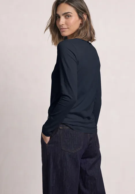 Frau im Profil mit dunkelblauem Langarmshirt und Jeans, Blick über die Schulter vor schlichtem Hintergrund.