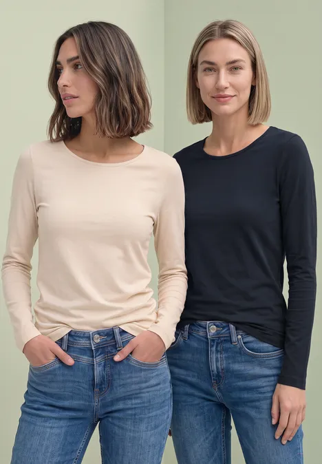 Zwei Frauen in Jeans und langärmligen Oberteilen (beige und schwarz) vor grünem Hintergrund.