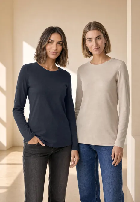 Twee vrouwen staan naast elkaar in een lichte ruimte, gekleed in effen lange mouwen shirts en spijkerbroeken.