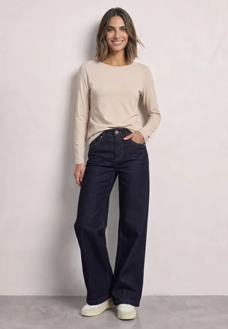 Frau steht vor heller Wand und trägt helles Langarmshirt mit dunkler, weiter Jeans