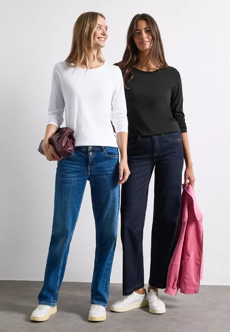 Zwei lächelnde Frauen in Jeans und Langarmshirts, jeweils mit Jacke oder Tasche in der Hand.