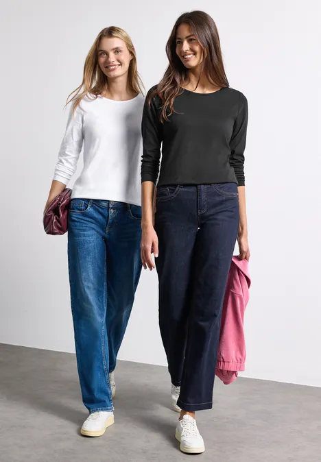 Zwei junge Frauen in lässigen Jeans und Langarmshirts posieren vor neutralem Hintergrund.