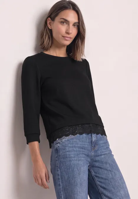 Frau in schwarzem Langarmshirt mit Spitzenbesatz und Jeans vor hellem Hintergrund.