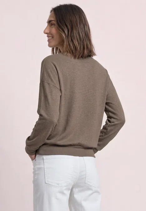 Femme de profil portant un pull marron et un pantalon blanc, mains dans les poches, sur fond clair.