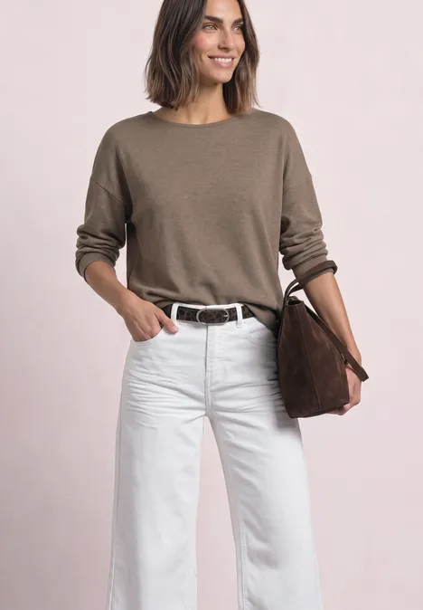 Une femme souriante porte un pull beige, un pantalon blanc et tient un sac marron.
