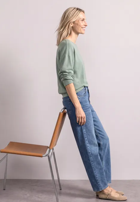 T-shirt à manches longues avec ourlet élastiqué eucalyptus green mel. Dame im grünen Pullover lehnt an Stuhl, trägt weite Jeans und flache Schuhe