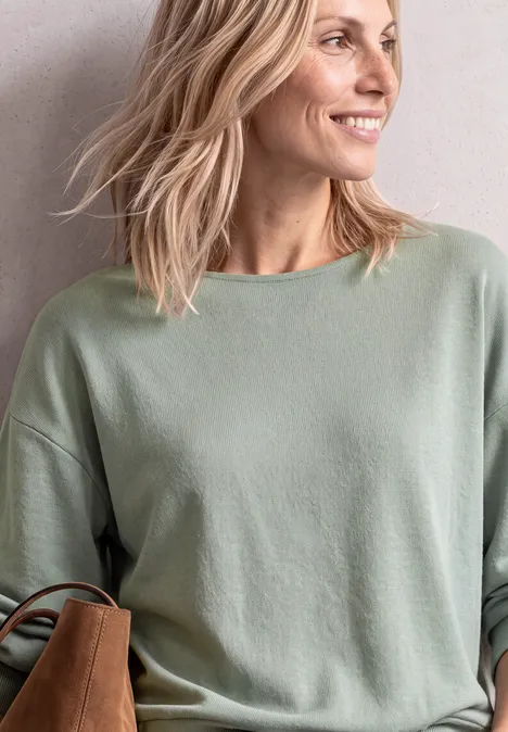 T-shirt à manches longues avec ourlet élastiqué eucalyptus green mel. Lächelnde Frau mit blondem Haar in grünem Pullover, seitlich blickend, mit Schultertasche.
