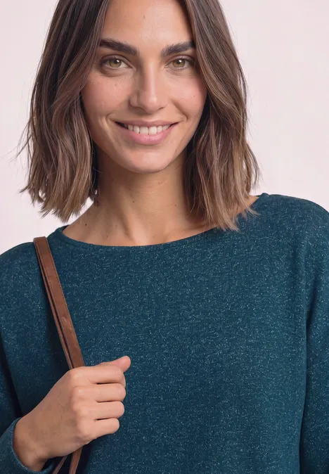Une femme souriante aux cheveux mi-longs portant un haut bleu-vert scintillant et un sac à bandoulière