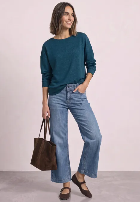 Femme en pull turquoise et jean large, portant un sac marron. Elle sourit légèrement.