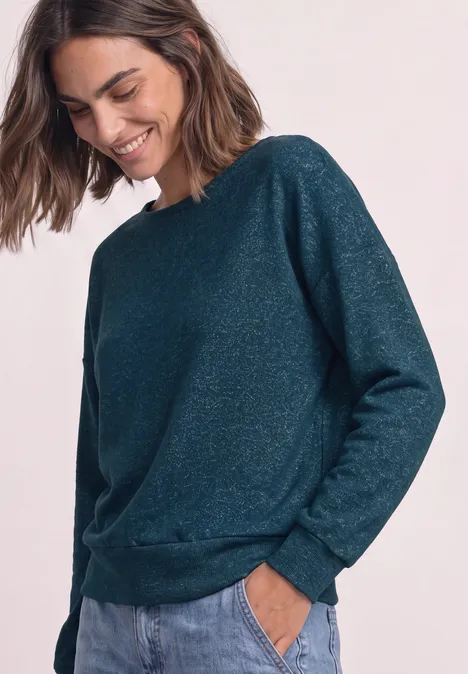 Femme souriante portant un pull vert foncé légèrement pailleté, manches longues et poches.