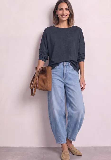 Femme en pull gris, jean ample, mocassins beiges et sac marron, posant devant un fond clair.