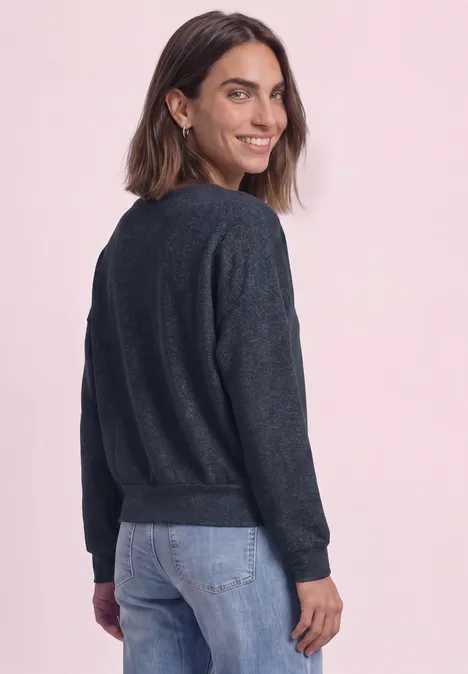 Femme souriante en pull bleu foncé et jean, de profil, regardant par-dessus l'épaule.