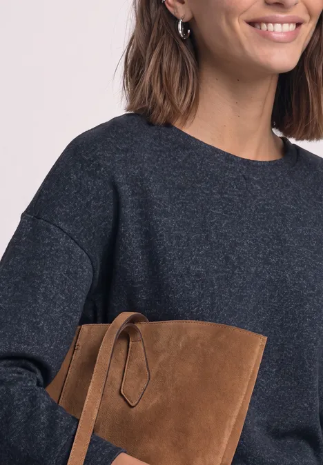 Femme en pull gris foncé souriante tenant une pochette en daim marron.