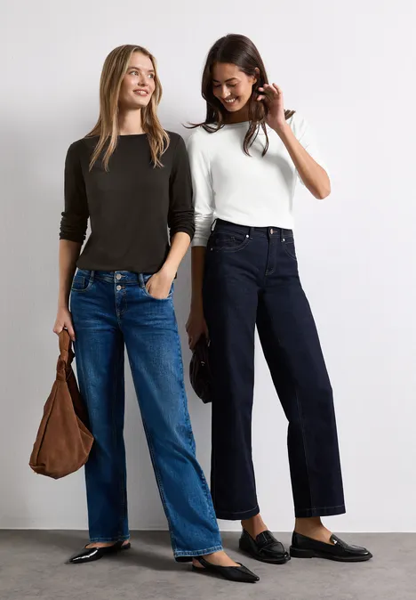 Zwei lächelnde Frauen in Jeans und schlichten Oberteilen stehen nebeneinander, eine trägt eine Tasche.
