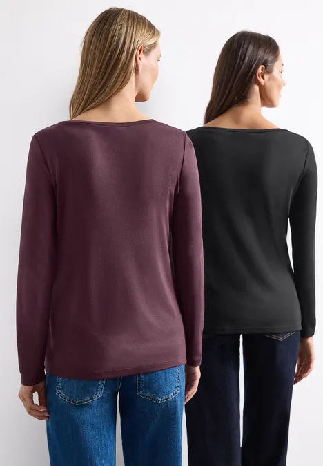 Zwei Frauen stehen mit dem Rücken zur Kamera, tragen Jeans und enge, langärmlige Oberteile in Schwarz und Bordeaux.