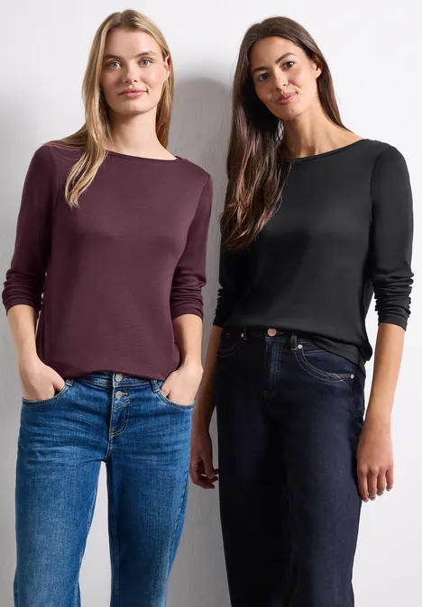 Zwei Frauen vor weißem Hintergrund: eine in einem burgunderfarbenen Top und Jeans, die andere in einem schwarzen Top.