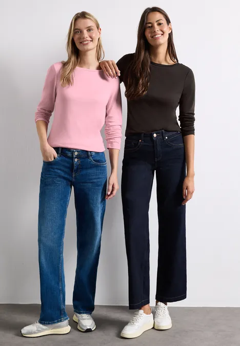 Zwei Frauen in lässigen Langarmshirts und weitem Jeans, lächelnd vor neutralem Hintergrund.