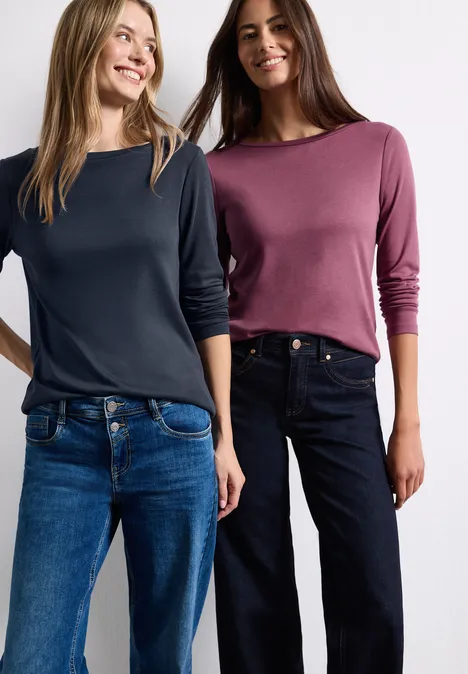 Zwei Frauen in lässigen Langarmshirts und Jeans stehen lächelnd nebeneinander.