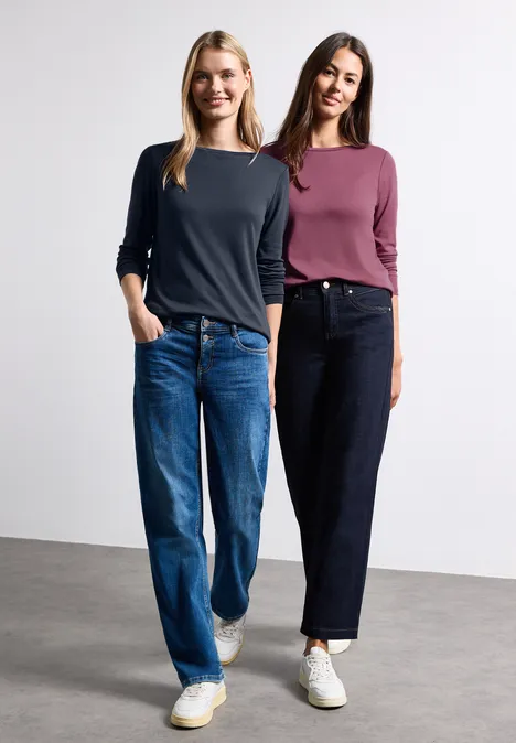 Zwei Frauen stehen nebeneinander in Jeans und lässigen Langarmshirts, tragen weiße Sneaker.