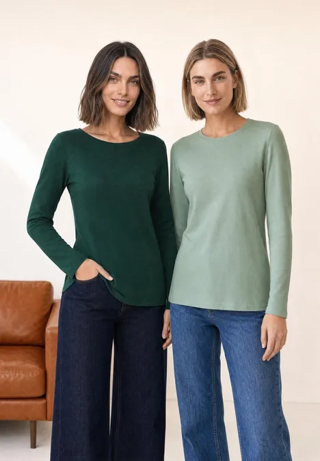 Zwei Frauen in grünen Langarmshirts und Jeans posieren nebeneinander vor einem hellen Hintergrund.