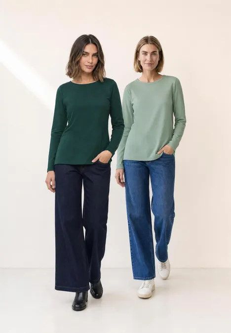 Zwei Frauen in grünen Pullovern und Jeans stehen nebeneinander vor neutralem Hintergrund.