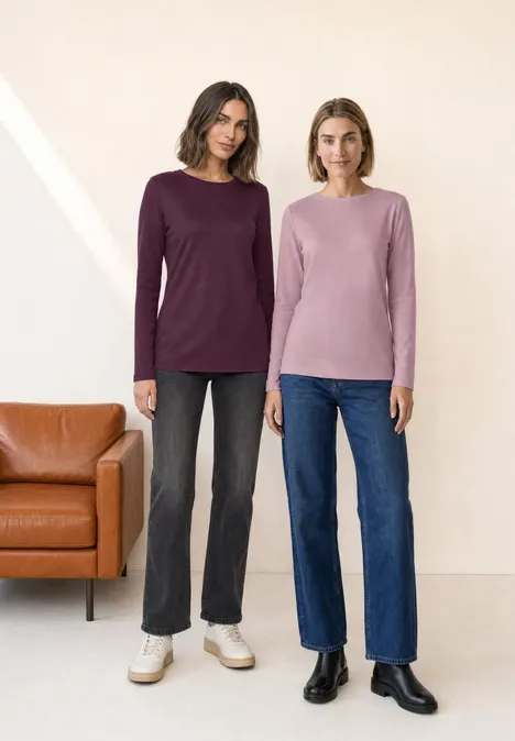 Twee vrouwen in truien en jeans staan voor een lichte muur naast een leren bank.
