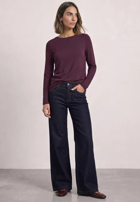 Frau mit langärmeligem, weinrotem Oberteil und weiten Jeans steht vor hellem Hintergrund.