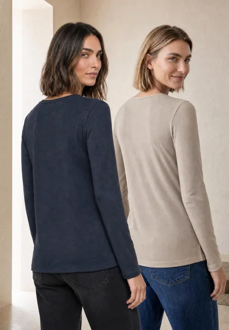 Zwei Frauen in Jeans und langärmligen Oberteilen (Marineblau und Beige) stehen nebeneinander und lächeln.