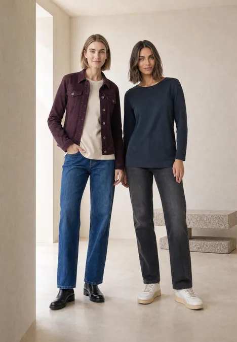 Zwei Frauen in minimalistischer Umgebung, tragen Jeans, Pullover und Jacke in neutralen Farben.