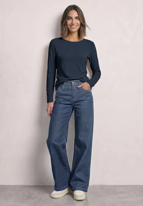 Eine Frau trägt ein langärmeliges dunkelblaues Shirt, weite Jeans und weiße Sneaker vor hellem Hintergrund.