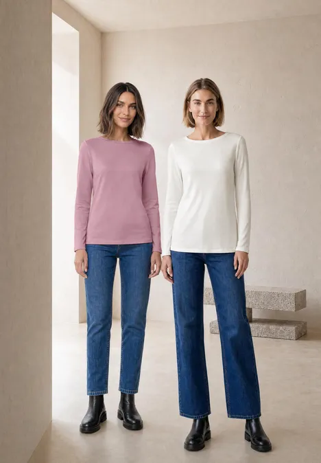 Twee vrouwen staan voor een lichte, minimalistische achtergrond en dragen jeans met lange mouwen.