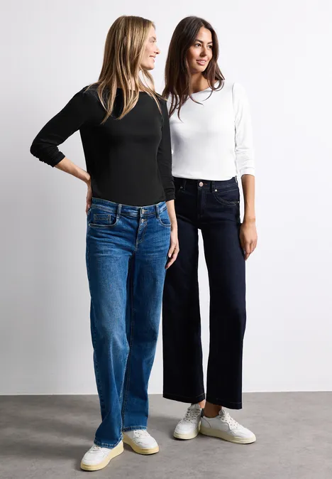 Zwei junge Frauen stehen nebeneinander, tragen lässige Jeans, langärmlige Oberteile und weiße Sneaker.