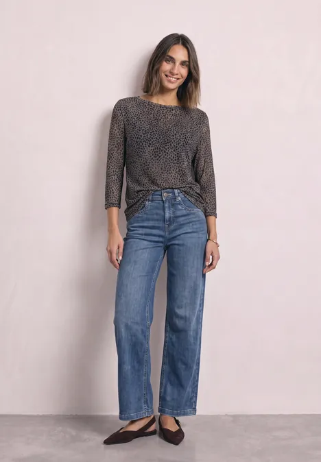 Frau mit schimmerndem Langarmshirt, weitem Jeans und schwarzen Ballerinas vor hellem Hintergrund.
