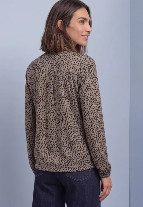 Frau von der Seite, trägt beige-braune Bluse mit schwarzem Leoprint und dunkler Hose vor schlichtem Hintergrund.